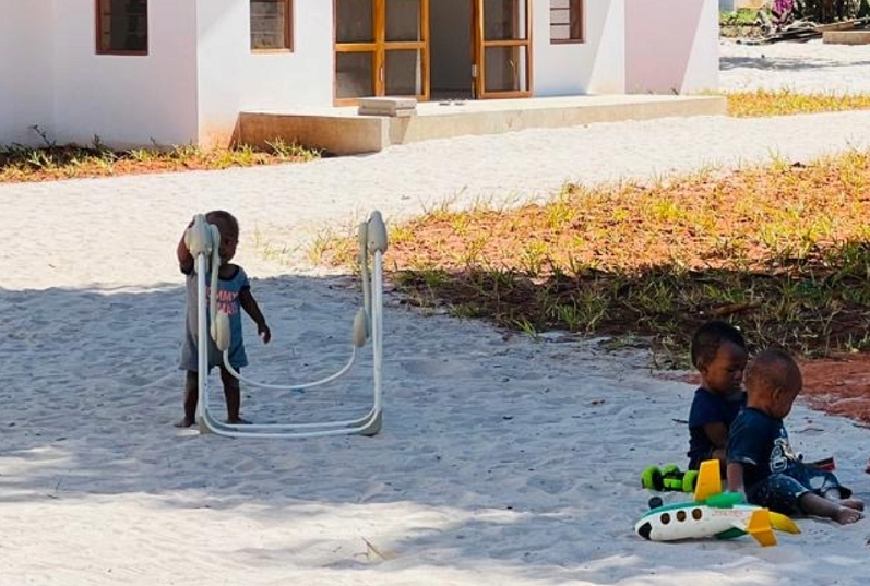 Two children are playing on the floor in front of a white building, while another child is pulling on a toy.