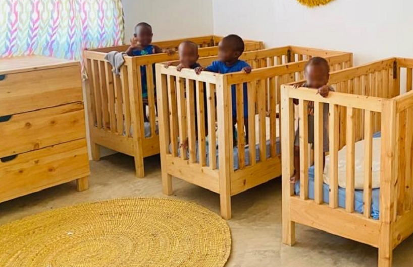 Three children are standing in wooden cribs in a bright room with colorful curtains and a round rug.