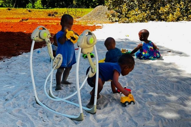 Children are playing in the sand while another child is busy with a toy. The surroundings are filled with greenery and a clear sky.
