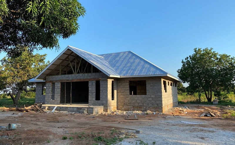 A half-finished stone house with a gabled roof, surrounded by trees and unpaved ground.