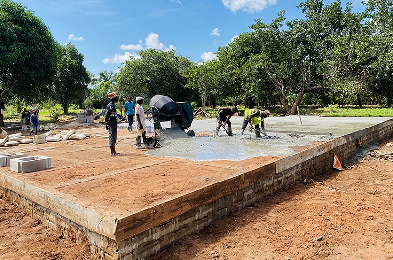 Workers are pouring concrete at a construction site to create the foundation of a building, surrounded by trees and natural scenery.