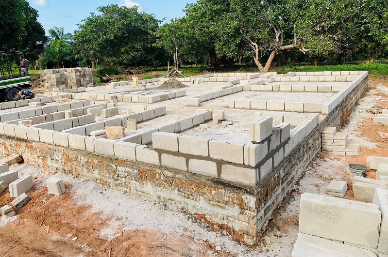 A building plot with a partially constructed wall made of concrete blocks, surrounded by trees and unpaved ground.
