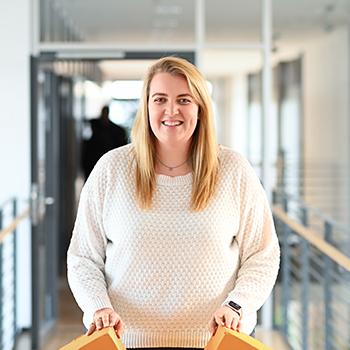 A smiling woman with long blonde hair is wearing a white sweater and holding orange boxes in a modern interior.
