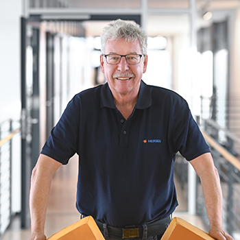 A man with glasses and gray hair stands in a modern hallway, holding two orange objects in his hands.