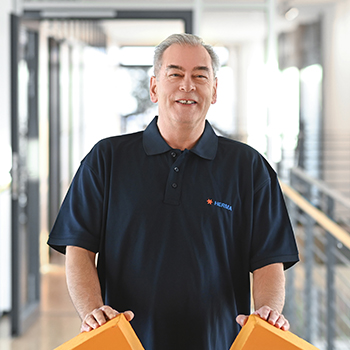 A man in a black polo shirt stands in a modern interior, holding two orange objects in his hands.