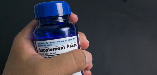 A hand is holding a blue bottle of dietary supplements, and the label displays the supplement facts.