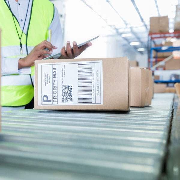A worker in a safety vest is checking packages on a conveyor belt while holding a smartphone in his hand.