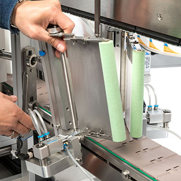 A hand adjusts a metal plate on a machine, while two green rollers are visible on the right side.