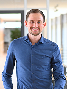 A man with short hair is wearing a blue shirt and smiling. He is standing in a modern office with large windows in the background.