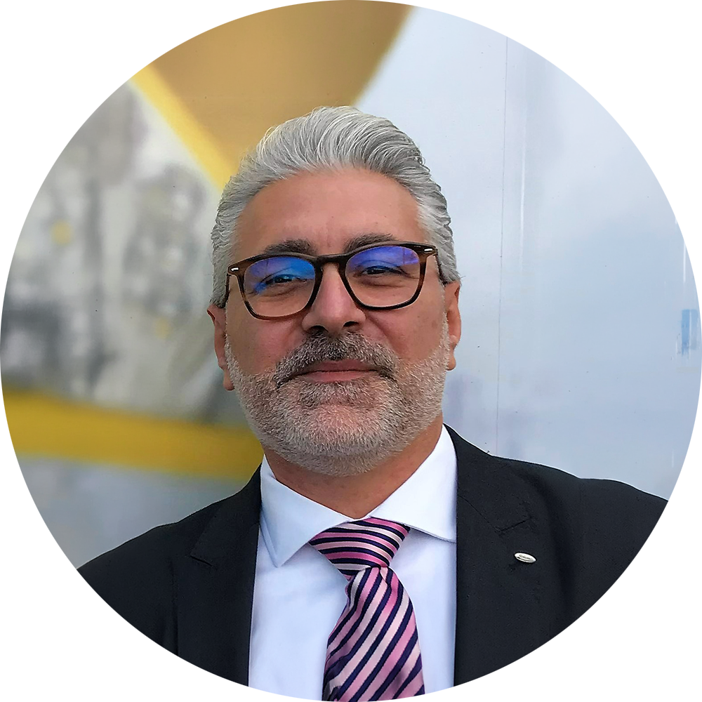 Man with gray hair and glasses, wearing a suit with a striped tie. Background is blurred and bright.