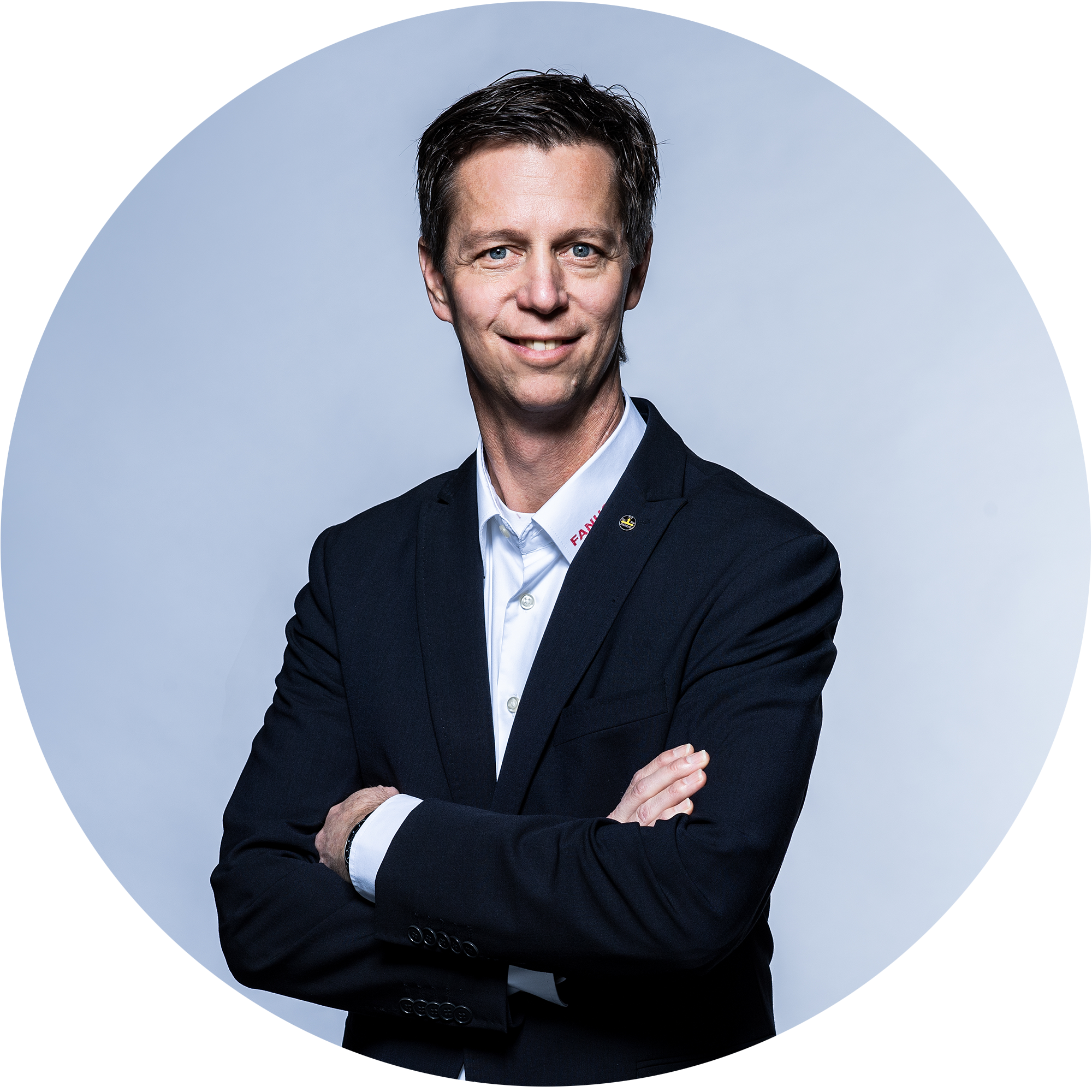 A man in a black suit with his arms crossed smiles at the camera. The background is bright and neutral.