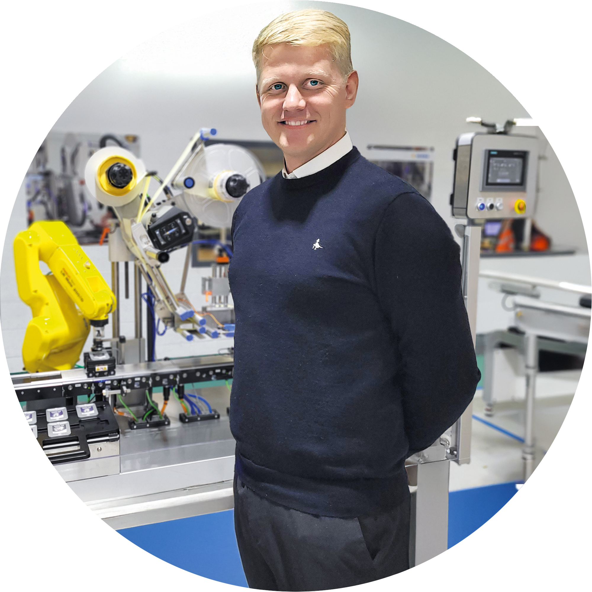 A man in a black sweater stands next to an automated production line with robots and technical equipment.