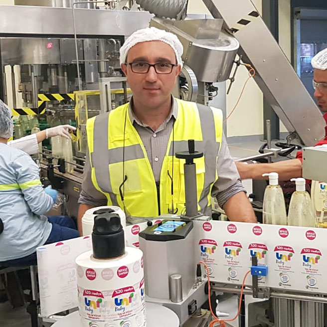 A man in safety gear and a hairnet stands in a production facility while employees fill bottles. Machines are running in the background.
