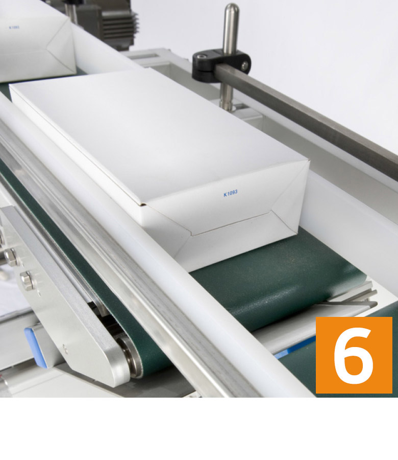 A conveyor belt transports several white packages, while a green surface is visible. An orange number 6 can be seen in the corner.