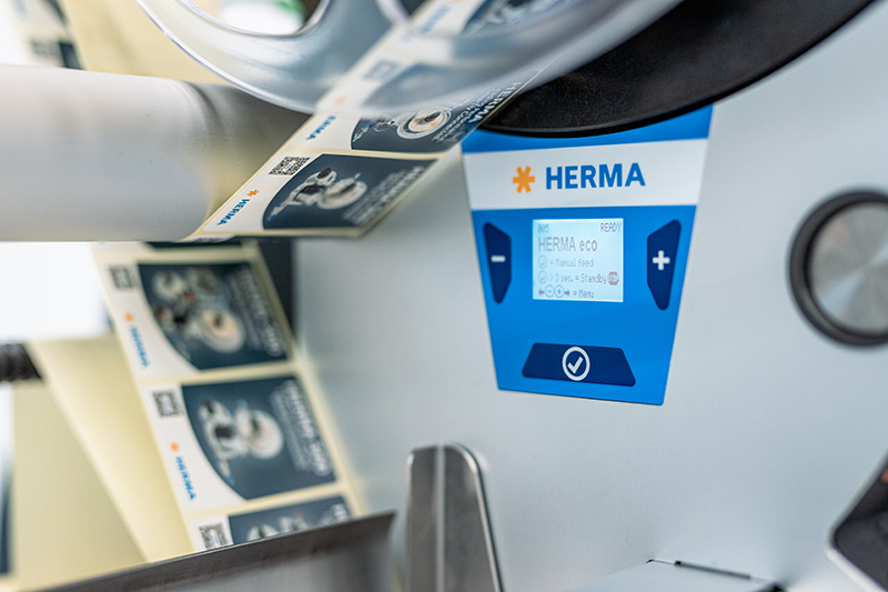 A label printer with a display showing the brand HERMA. Labels are dispensed on a roll.