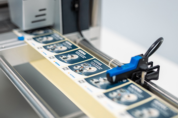 A printing machine processes labels with technical illustrations and text. The labels are arranged on a roll.