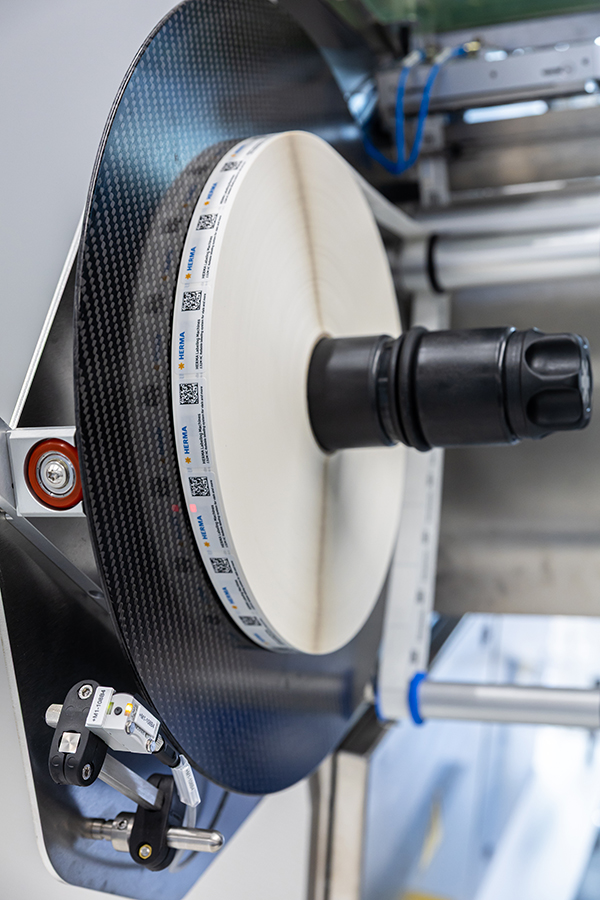 A large, round carbon reel with a central black holder, surrounded by technical components and sensors.