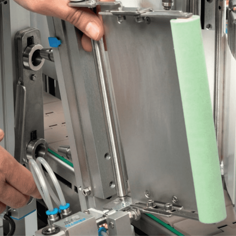 A hand adjusts a mechanical device with metal parts and a green component. The surroundings feature industrial equipment.