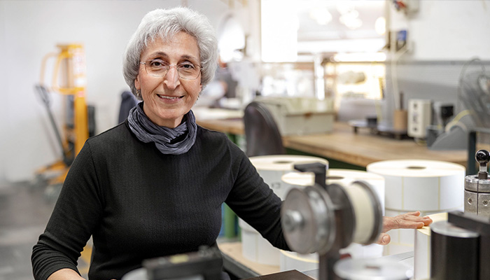 An older woman with gray hair sits at a table in a workshop, surrounded by spools and machines. She smiles warmly.