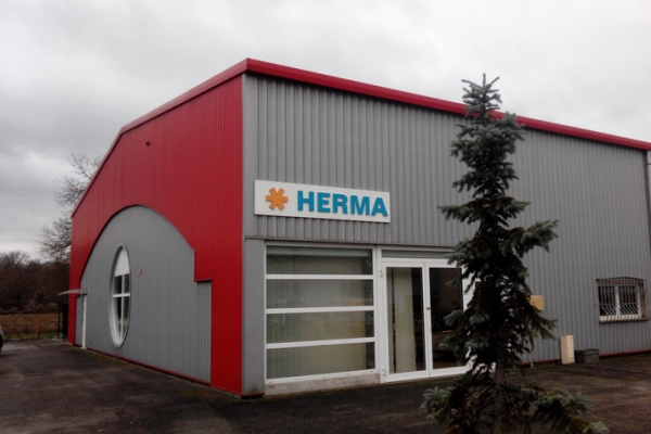 Facade of a modern building with a red roof and gray walls, labeled with 'HERMA.' A tree stands in the foreground.
