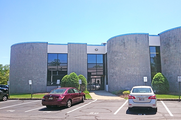 Modern buildings with gray facades, large windows, and two parked cars in the foreground. Green shrubs flank the entrance.