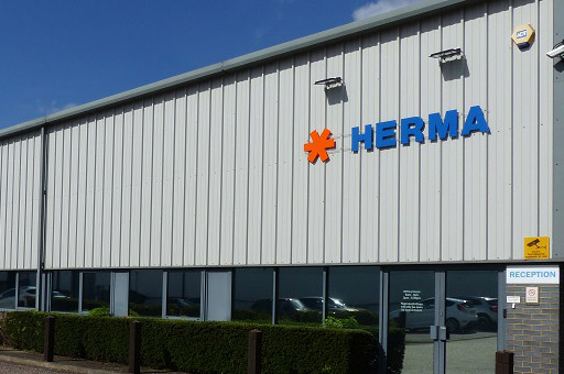 Facade of a modern building featuring the inscription "HERMA" in blue lettering and an orange symbol. Reception area visible.