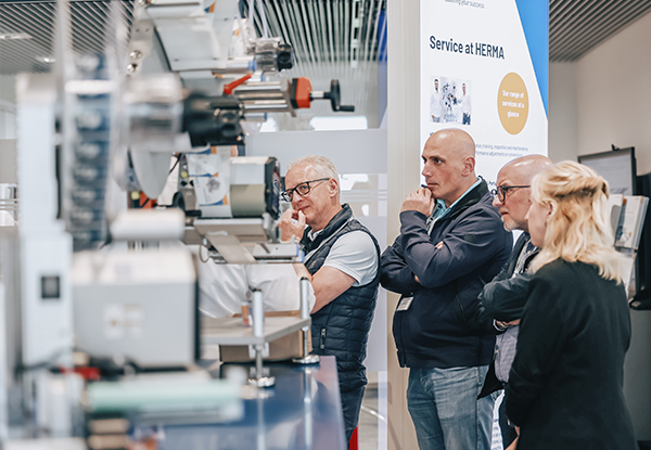 Four people are attentively watching a machine presentation at a trade show, while information about the service at HERMA is being displayed.
