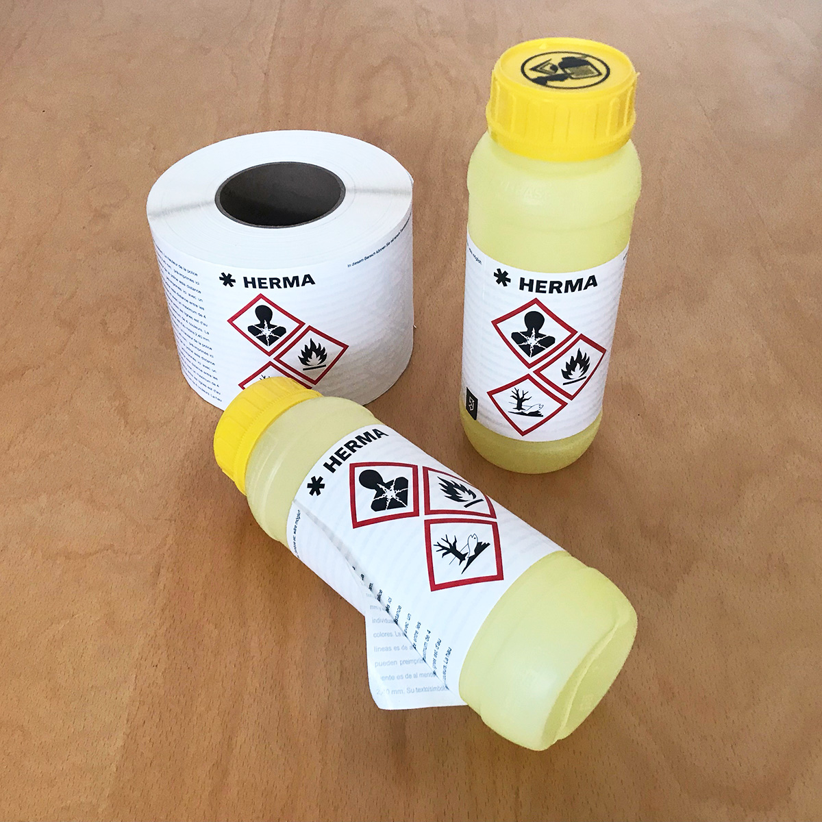 Application for wrap-around labels: Two yellow bottles with wrap-around labels that feature a hazardous material warning, and a roll of HERMA wrap-around labels on a wooden surface. Wrap-around labels are the ideal solution for the extensive and stringent labeling requirements set forth by the new CLP regulation.