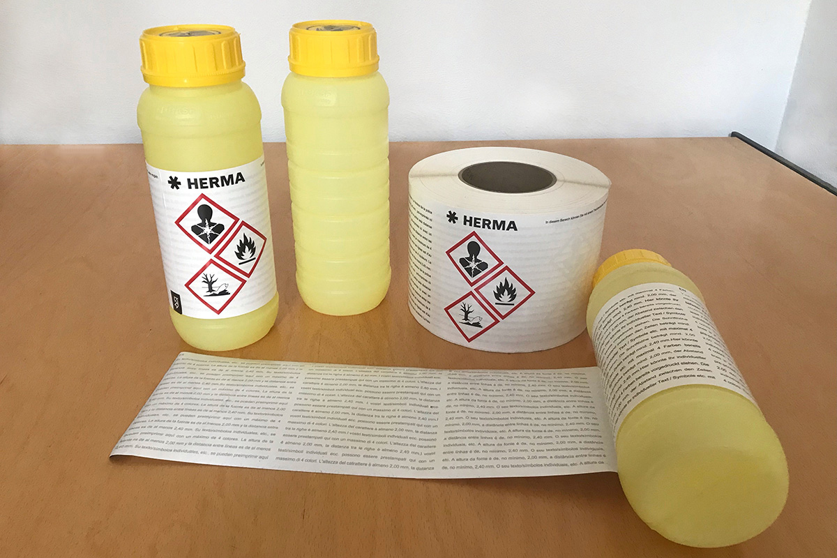 Two yellow bottles with hazard symbols and a roll of labels on a table. The labels contain safety information.