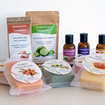Labels on various cosmetic products: Doypacks with bath milk and bath powder, brown glass bottles with bath aroma, as well as plant oil soaps with round labels in clear bags, set against a light background.