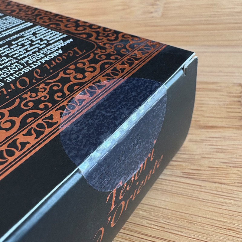 Cosmetic solutions: Black packaging with decorative orange patterns and text. The flap of the packaging is sealed with a transparent closure label.