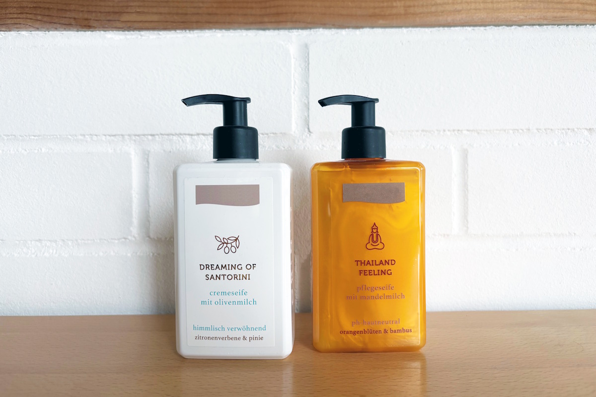 Two pump dispenser bottles of liquid soap: one white bottle with a white label and one orange bottle with a transparent film label in a no-label look, placed on a wooden surface in front of a white brick wall.