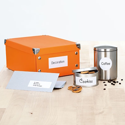 Printed universal labels on orange box, silver coffee container, tin with cookies and grey envelope on a wooden table.