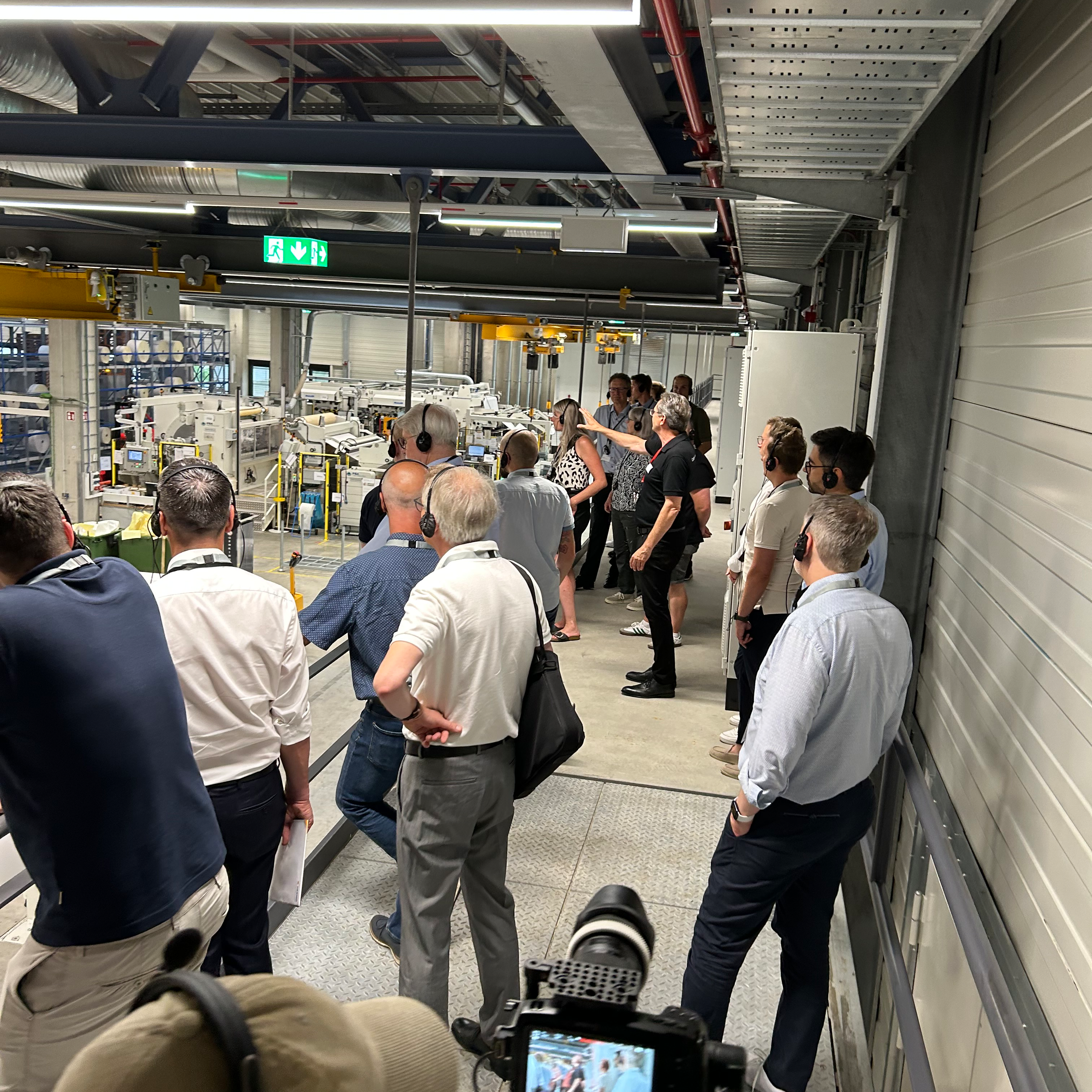 Group training at a production facility, participants observe machines and engage in discussions. Some individuals are taking notes or photographing.