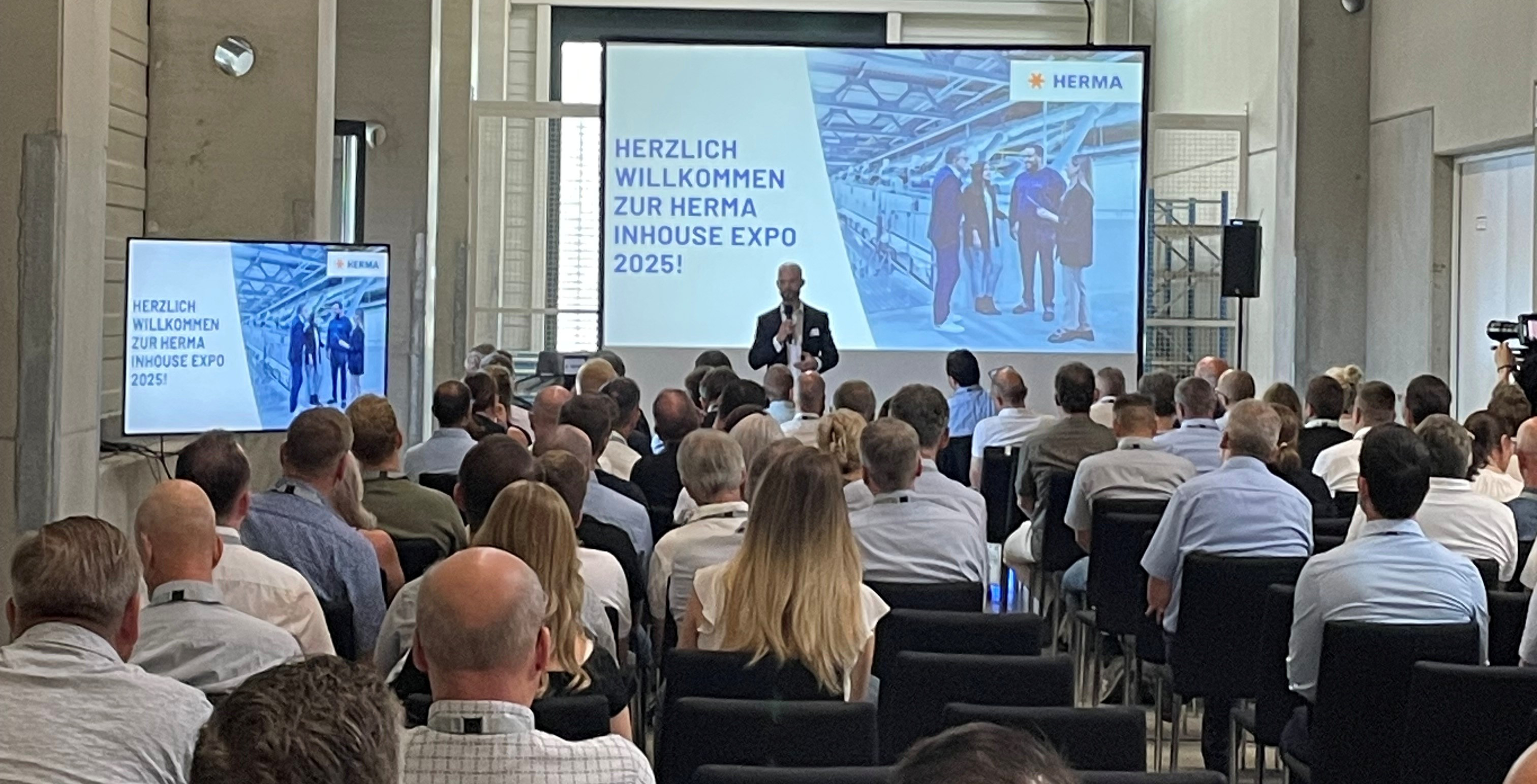 A speaker is presenting at the HERMA Inhouse Expo 2025 in front of an audience. In the background, a slide with a welcome message is visible.