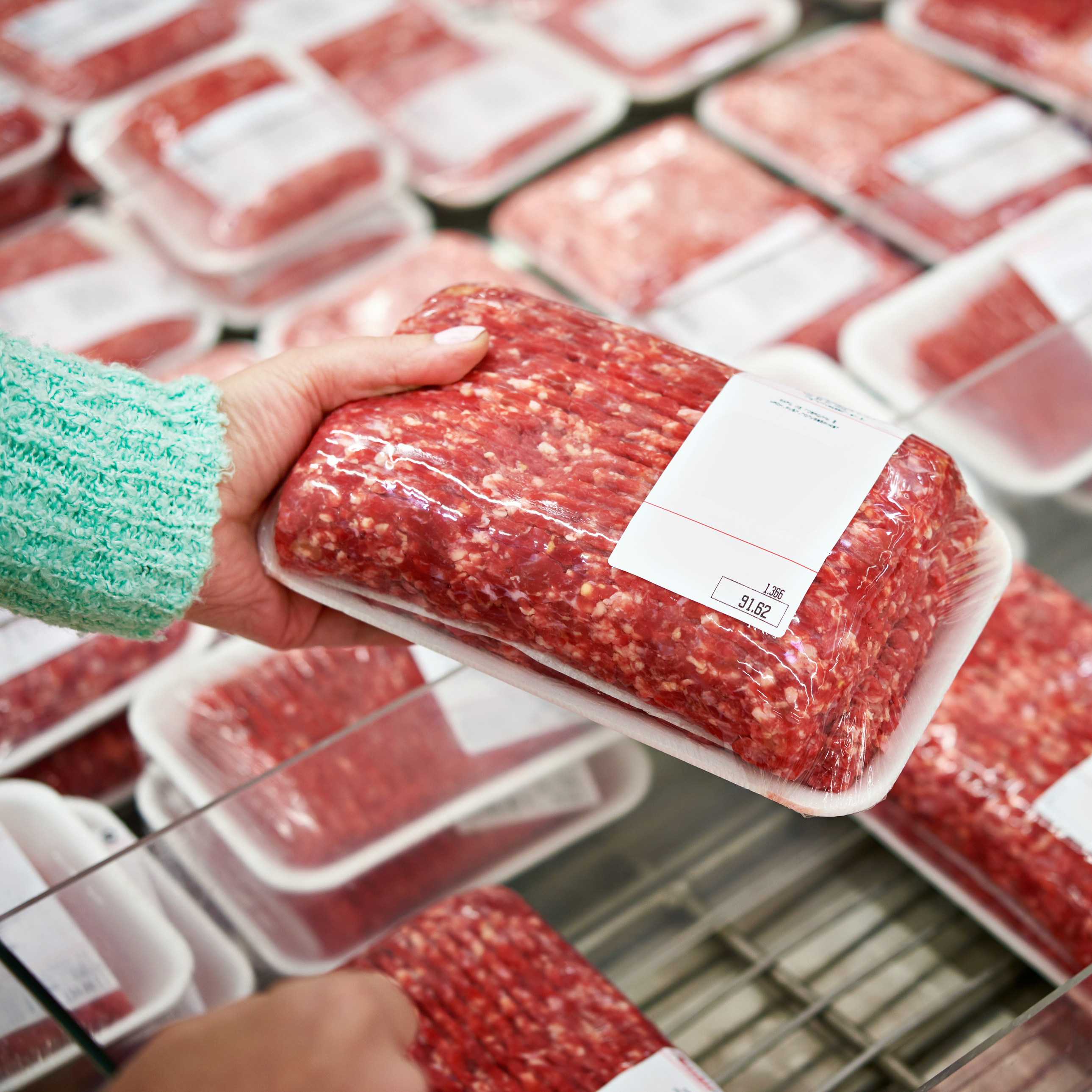 A hand is holding a package of ground meat, while in the background, more packaged ground meat products are visible.