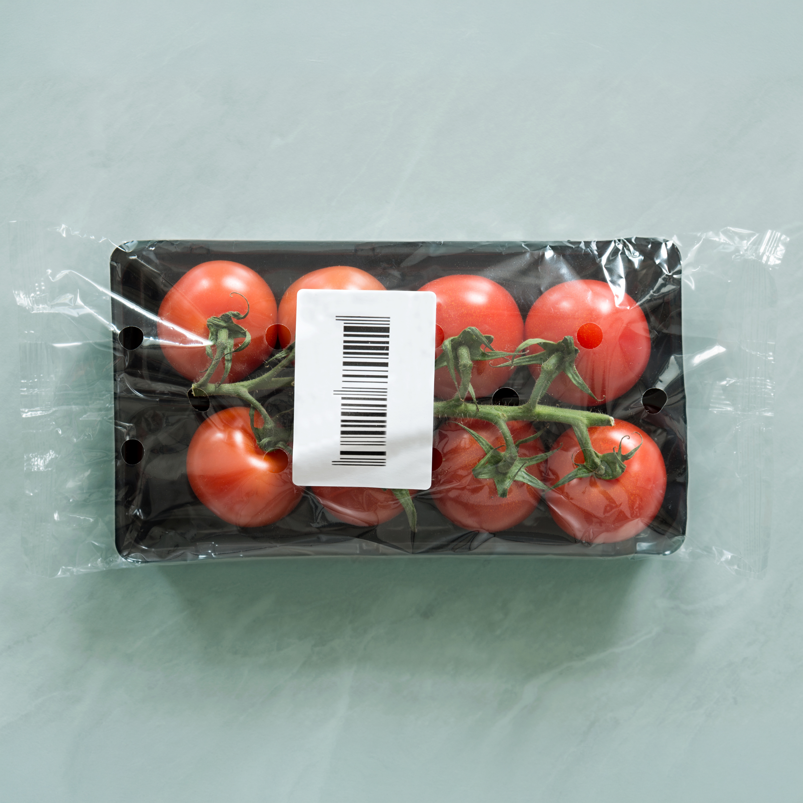 Six red tomatoes in a black packaging with a barcode. The tomatoes are attached by their stems.