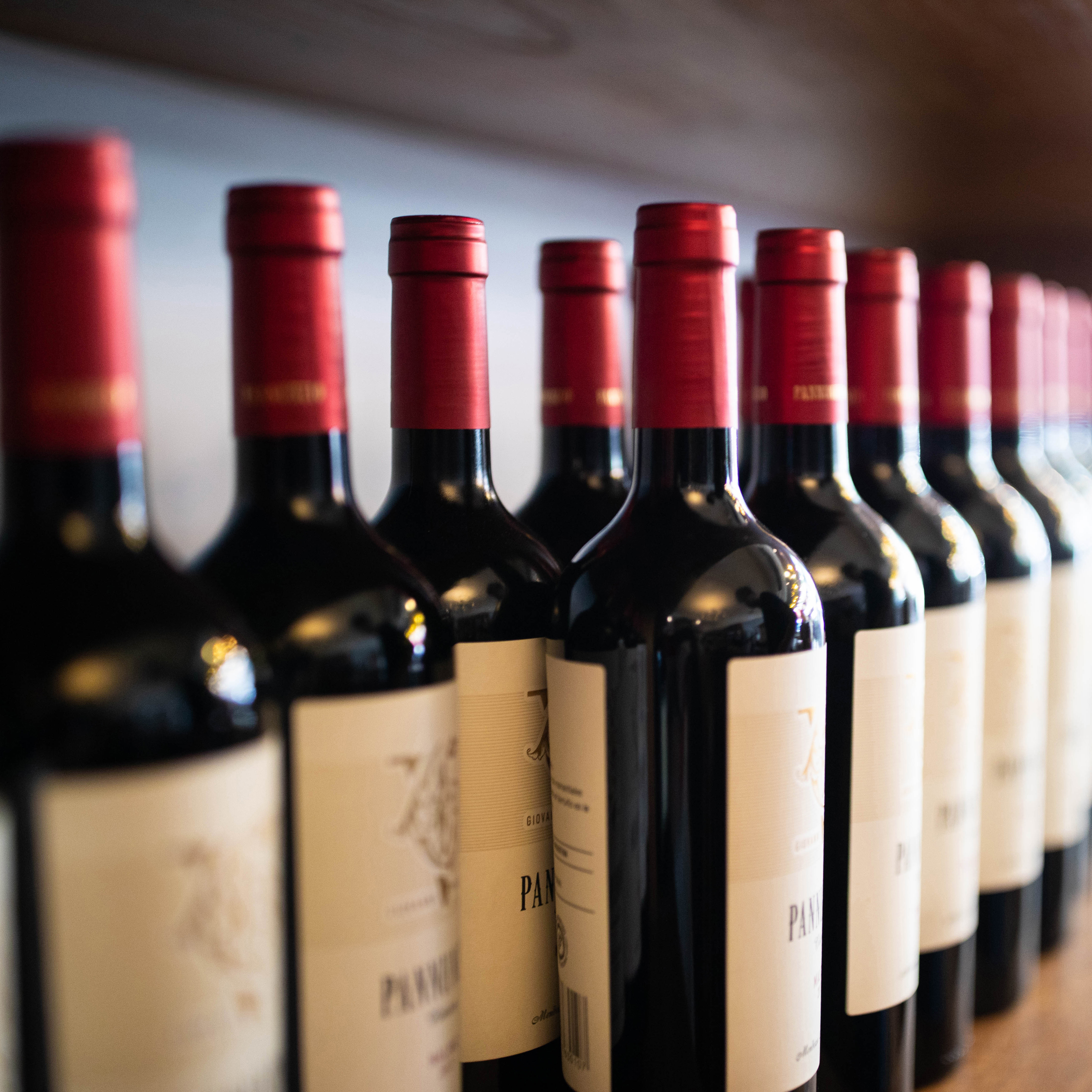 Row of red wine bottles with red caps, labeled and arranged on a shelf.
