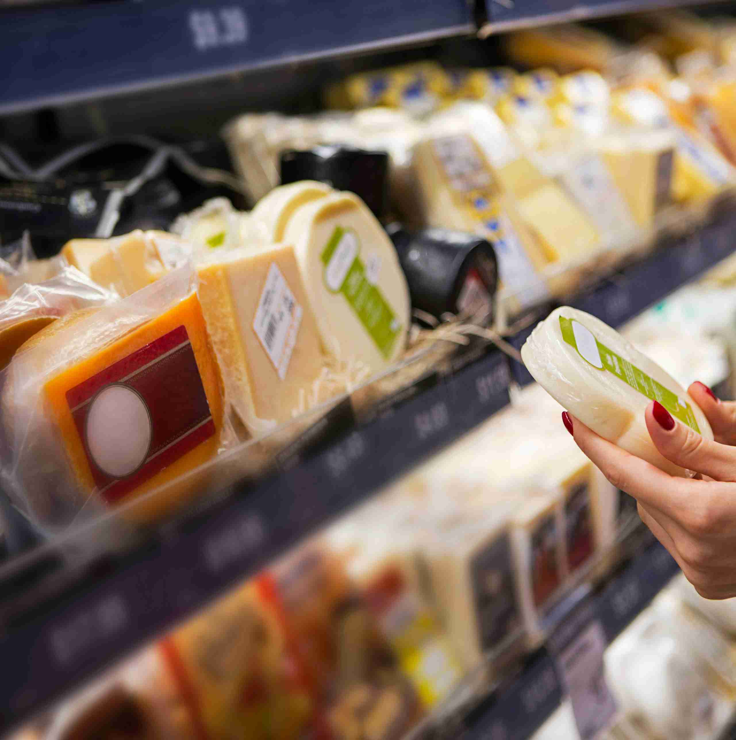 A hand holds a piece of cheese in front of a shelf displaying various types of cheese packaged for sale.