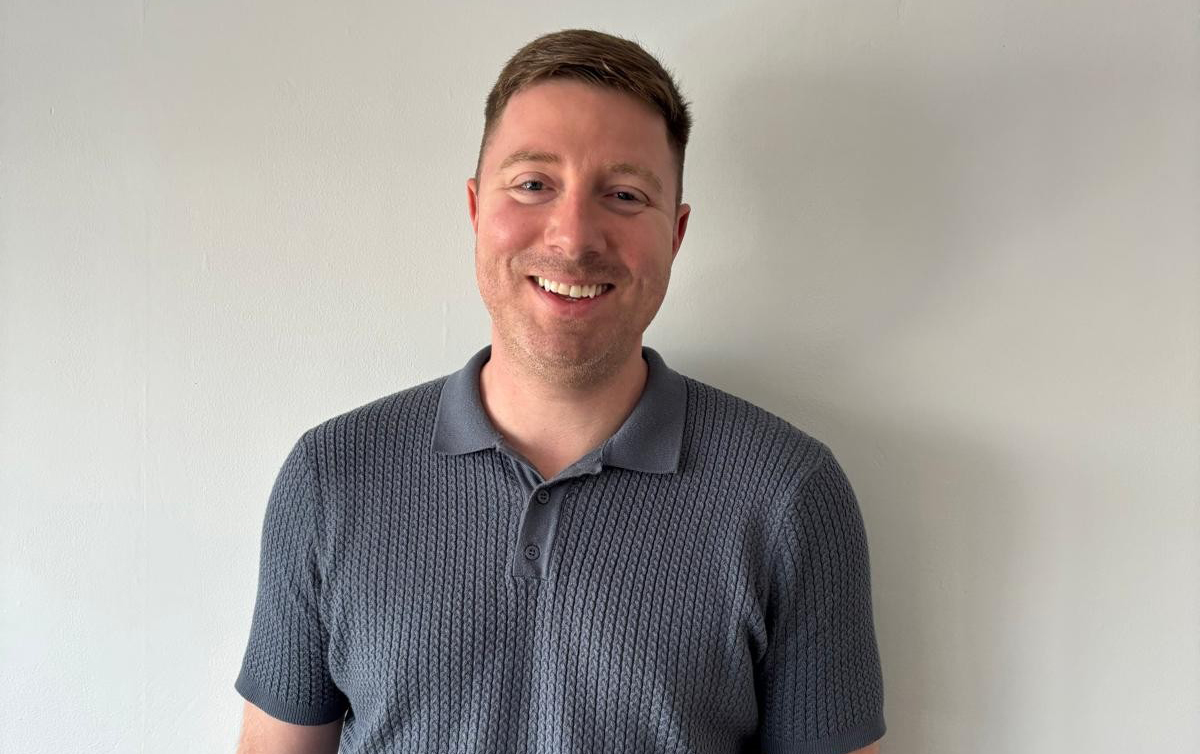A man with short brown hair is wearing a gray polo shirt and smiling in front of a neutral wall.