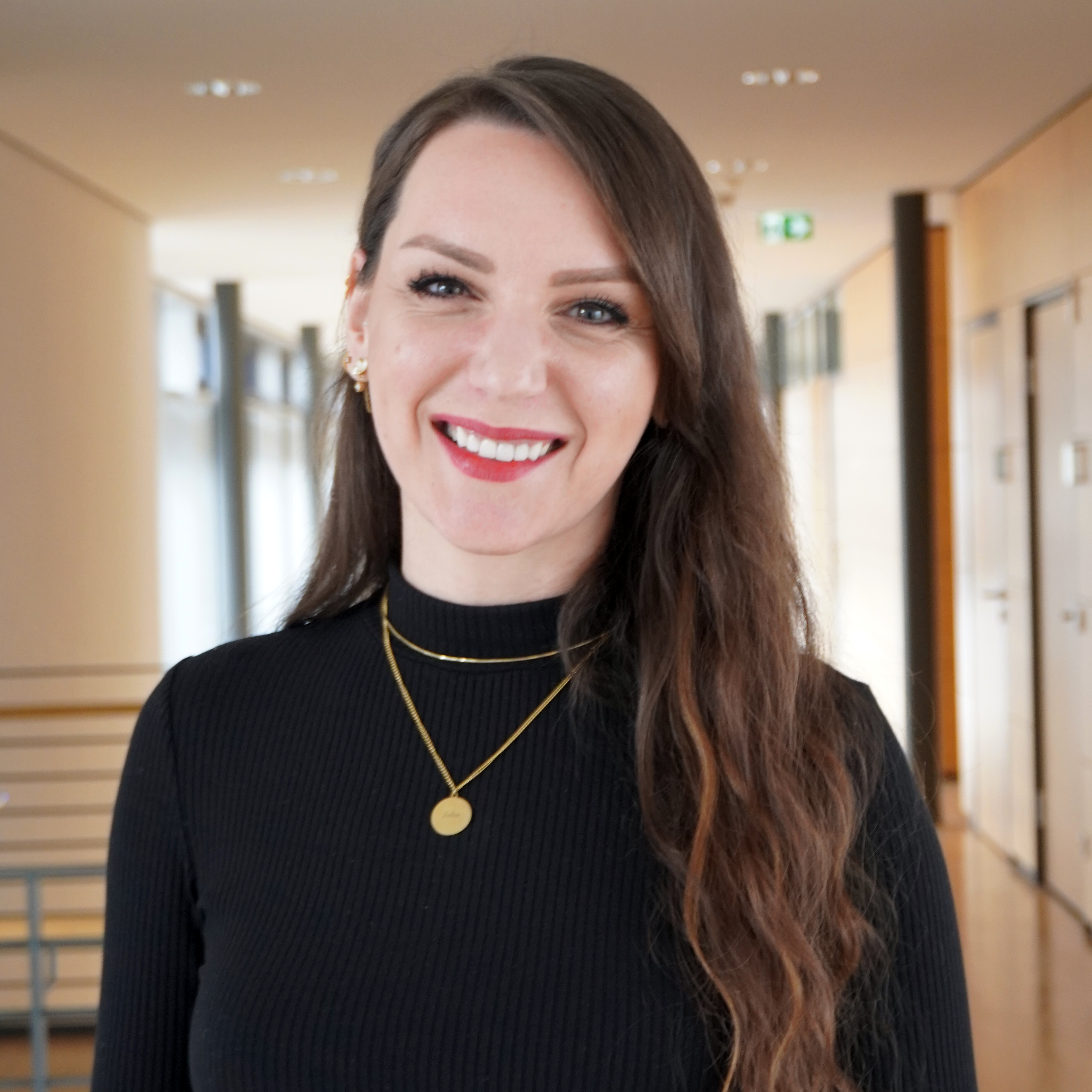 A woman with long, curly hair is wearing a black top and jewelry. She is smiling warmly in a modern interior.
