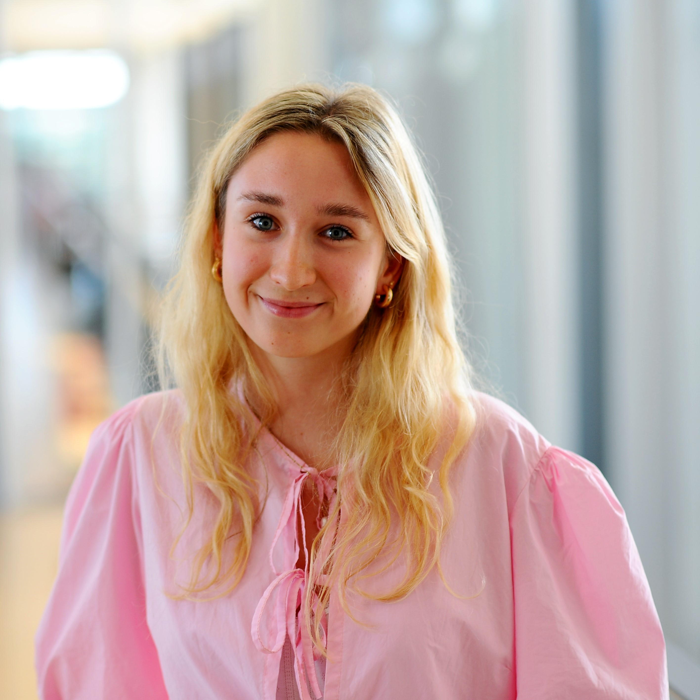 Young woman with long blonde hair and a pink top, smiling in a modern interior.