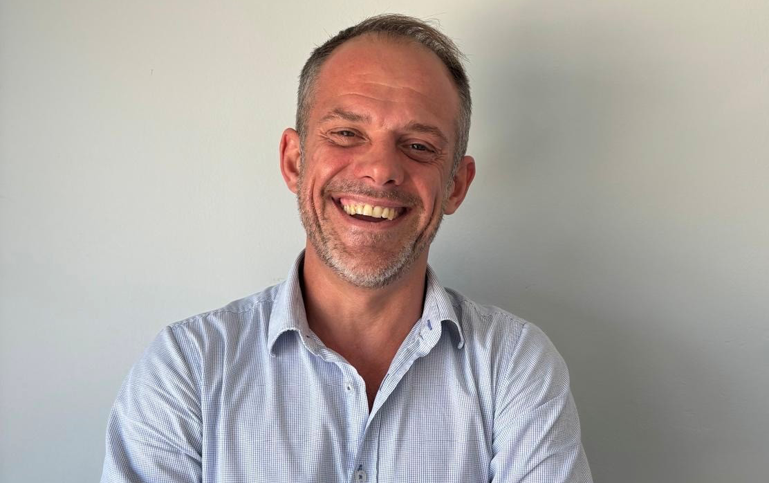 A man with short gray hair and a broad smile is wearing a light blue shirt and stands with his arms crossed in front of a neutral wall.
