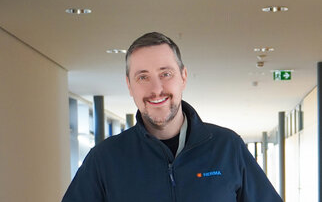 A man with short hair and a beard is wearing a dark jacket and smiling warmly in a modern hallway.