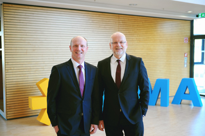 Two men in suits are smiling in front of large, colorful letters that spell out 'MMA' in a modern interior.