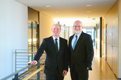 Two men in suits are standing in a modern hallway. The man on the left is wearing a patterned tie, while the one on the right has a beard and glasses.