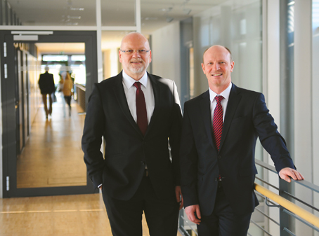 Two men in suits are standing in a modern hallway. The man on the left has glasses and a beard, while the one on the right is bald.
