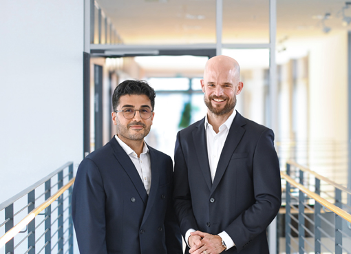 Two men in suits are standing in a modern office hallway, smiling at the camera. The background features bright, open architecture.