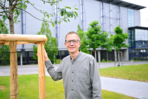 A man with glasses stands next to a young tree and a wooden frame in front of a modern building with large windows.