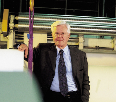A man in a suit stands by a machine in an industrial setting, wearing a neutral expression and having gray hair.